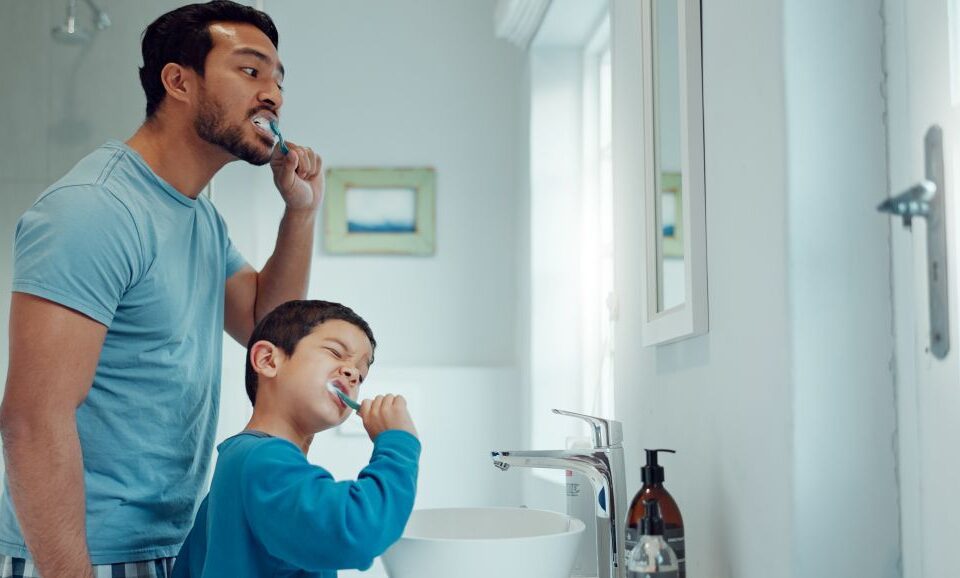 Man and child brush teeth together looking at mirror.